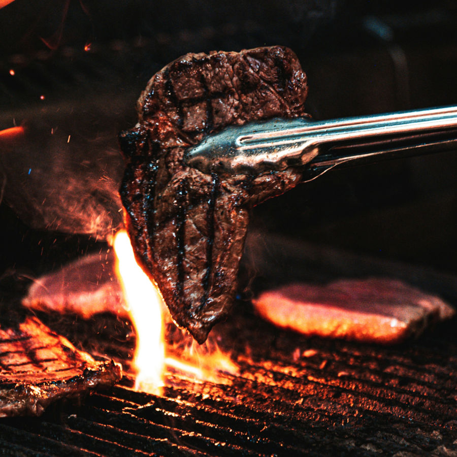 Ein Stückfleisch wird auf dem Grill mit einer Grillzange gewendet. Eine Flamme ist durch das Grillrost zu sehen.